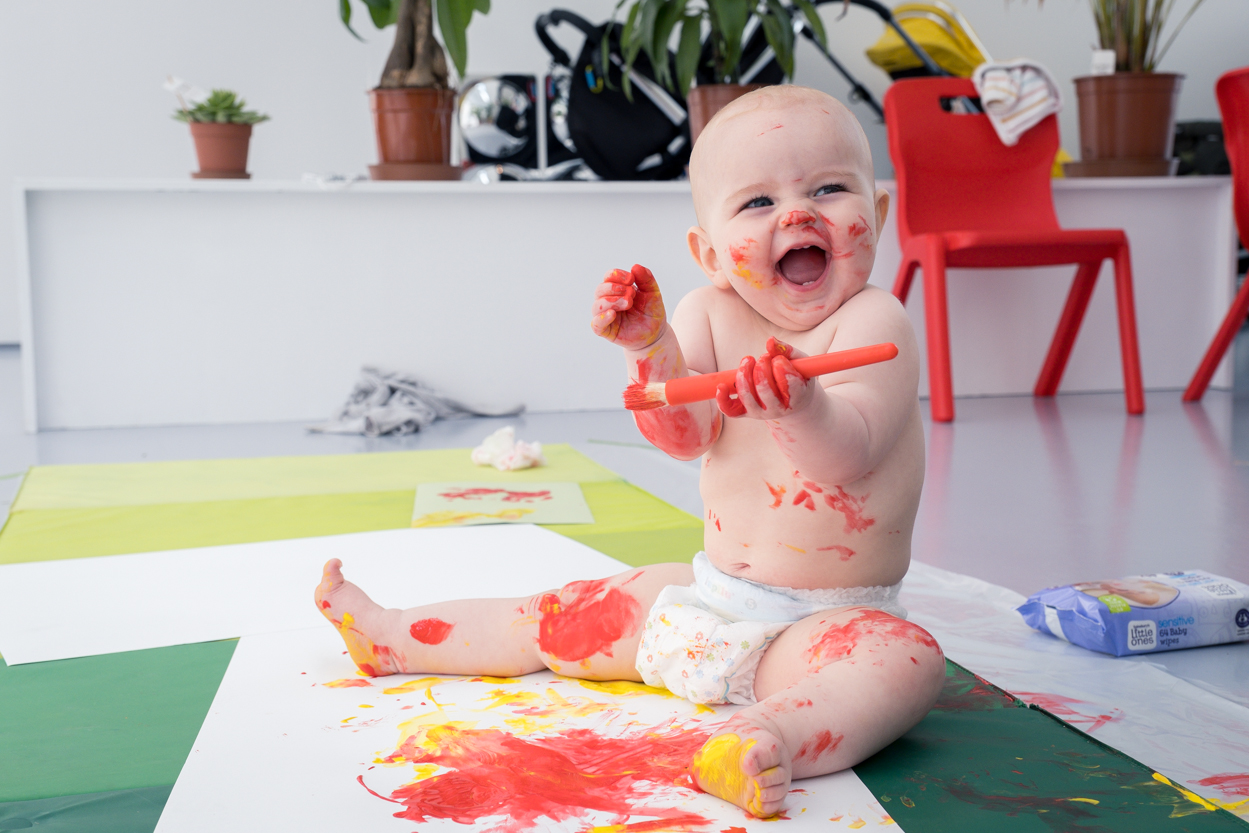 A baby holds a paintbrush and sits on white paper on the floow. It is covered in paint from its first painting attempts and seems to be very happy.