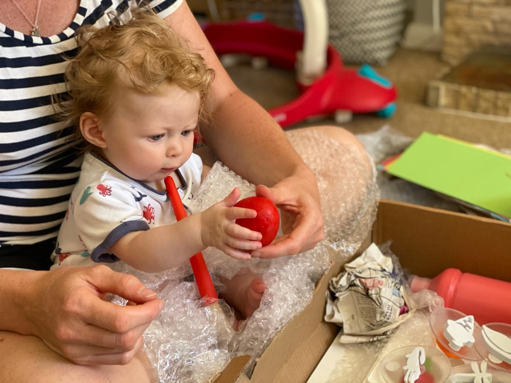 A toddler sits on an adults lap and is touching a red ball. Various other sensory objects are in front of them, e.g. bubblewrap.