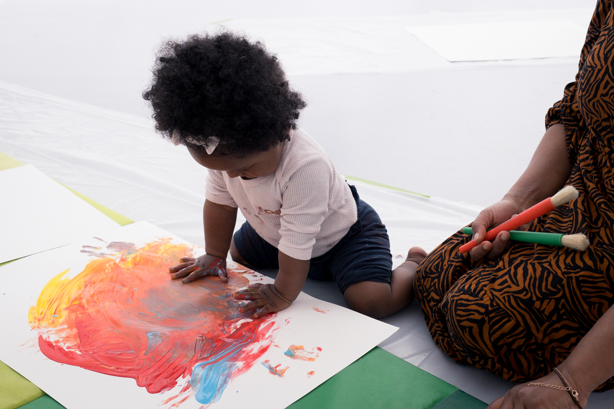 A toddler sat on the floor, painting with fingerpaint, hands full of blue, red and yellow paint.