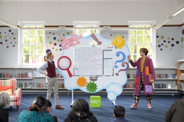 Two actors of Book Sleuths in a Bind are performing in a library. Between them, there is a big board with clues.