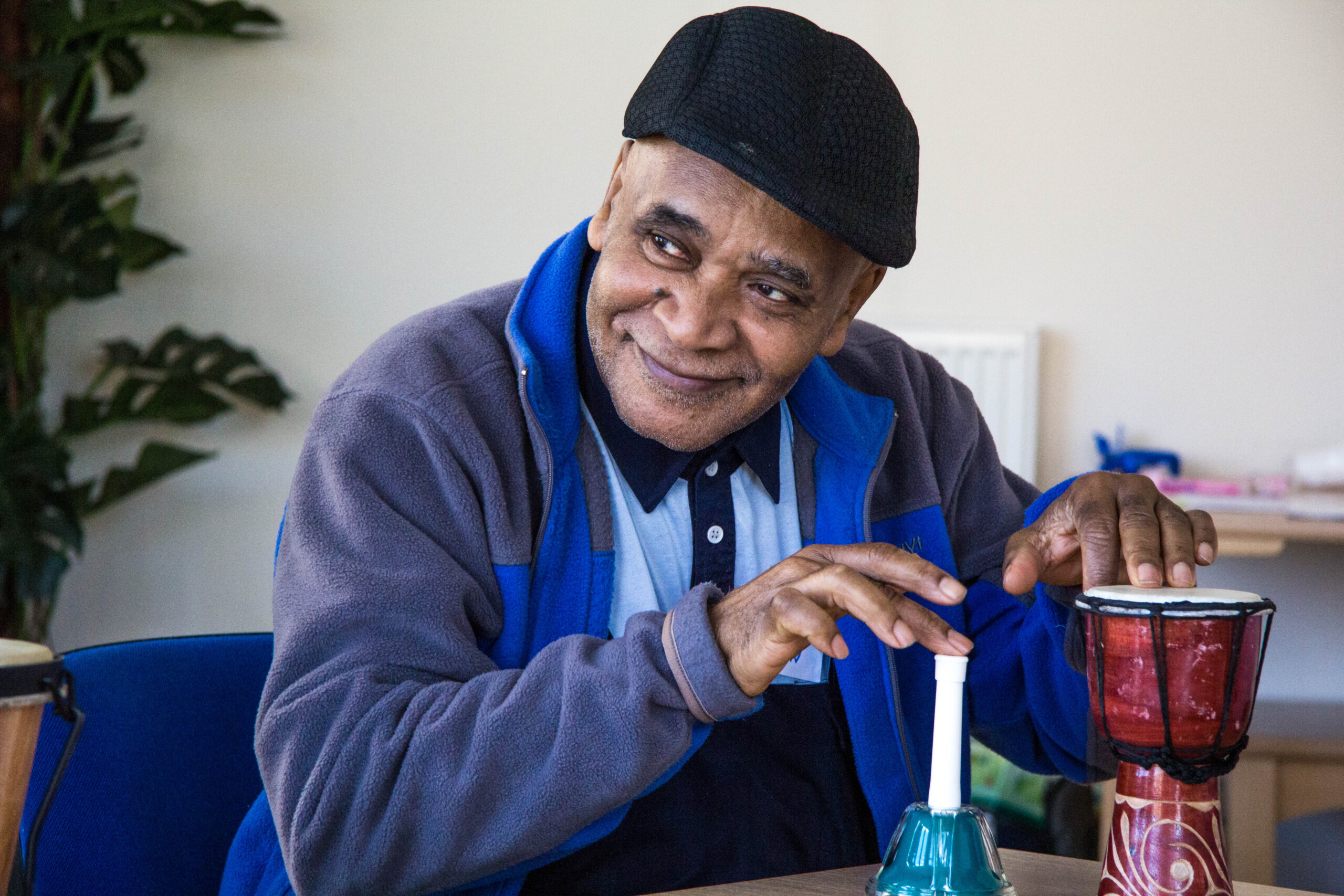 A Black man with a black cap, blue fleece and light blue polo t shirt sits at a table with one hand on a small drum and the other on a stick. There is a white wall behind in the room and a plant.