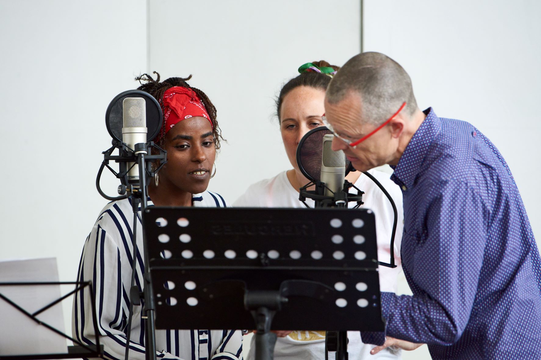 Three singers gathered around studio mics, looking at something on a music stand