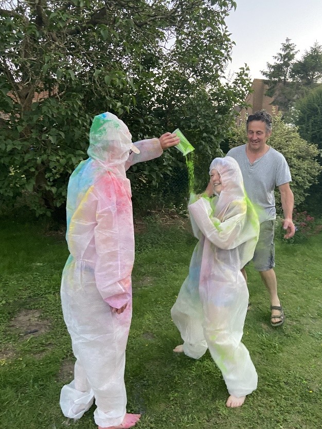 Three people, two of them wearing coveralls, are using the July sensory box to have an 'Outdoor Powder Paint Party': pouring colourful powder on one another.