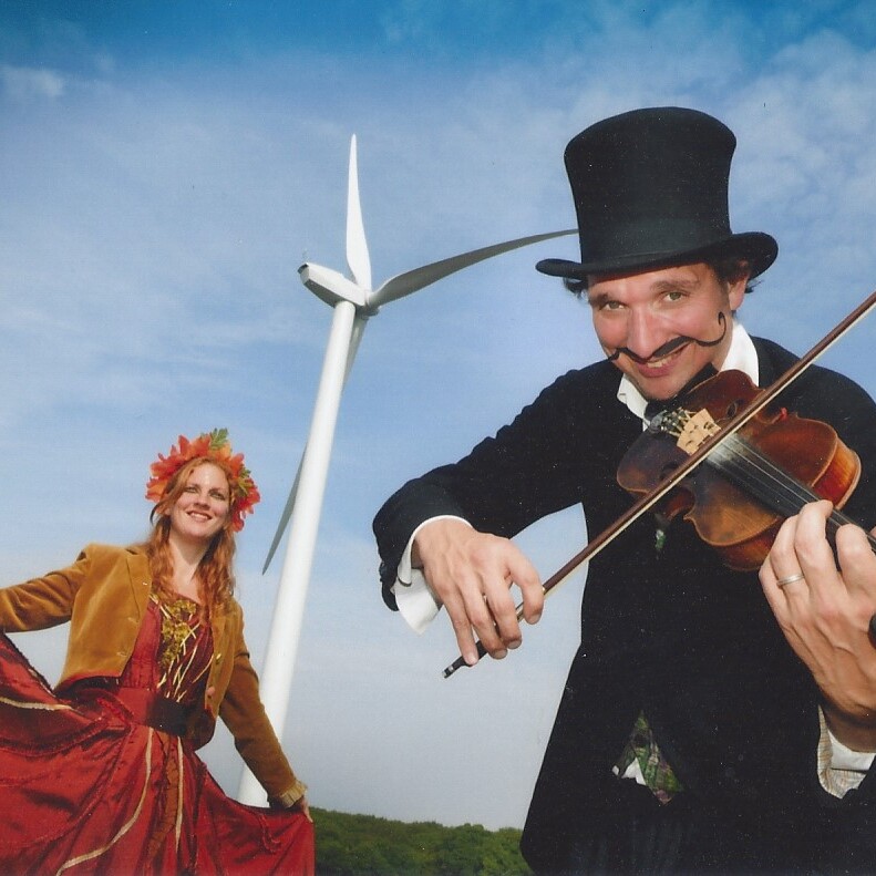 Two performers. The one on the left is wearing a rust-coloured dress and a leaf-crown. The one on the right is wearing a black top hat and playing the violin. In the background: a wind power station.