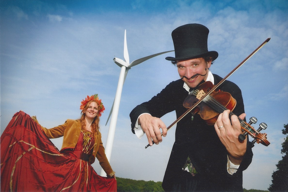 Two performers. The one on the left is wearing a rust-coloured dress and a leaf-crown. The one on the right is wearing a black top hat and playing the violin. In the background: a wind power station.