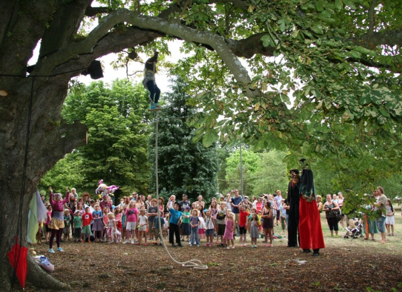 A person abseiling from a tree. Under the tree, there is an audience of mostly children. Performers on stilts stand on the side.