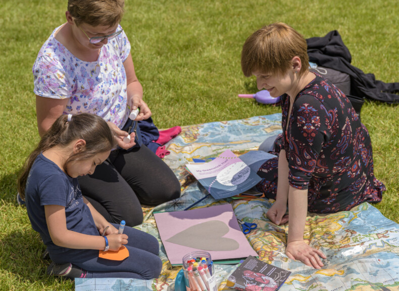 Two adults and a child engaging in arts and crafts together on a picnic blanket outside