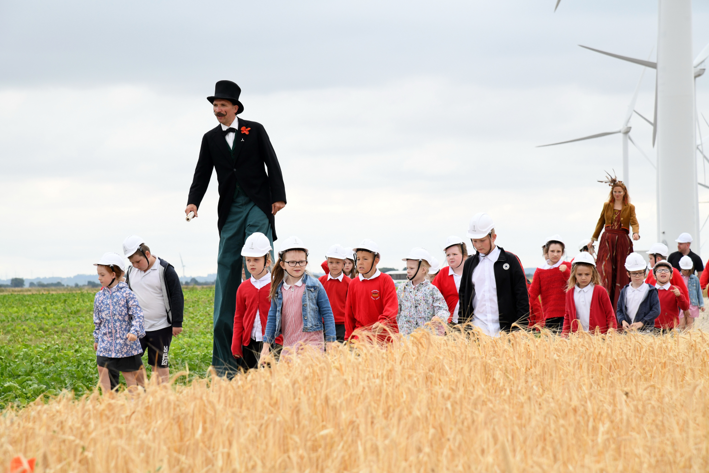 A Positive Imaginings workshop. Performers on stilts walk through the countryside together with children wearing white hardhats.