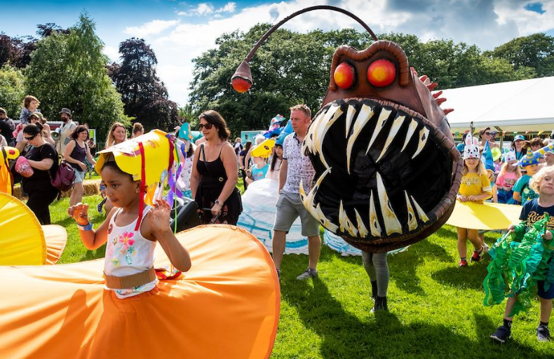 Costumed and children and adults perform outdoors at The Big Malarkey Festival. Notably, one person is dressed up as an anglerfish.