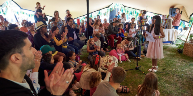 A tent at Walthamstow Garden Party, where a child wearing a pink dress is speaking into a microphone in front of a family audience.